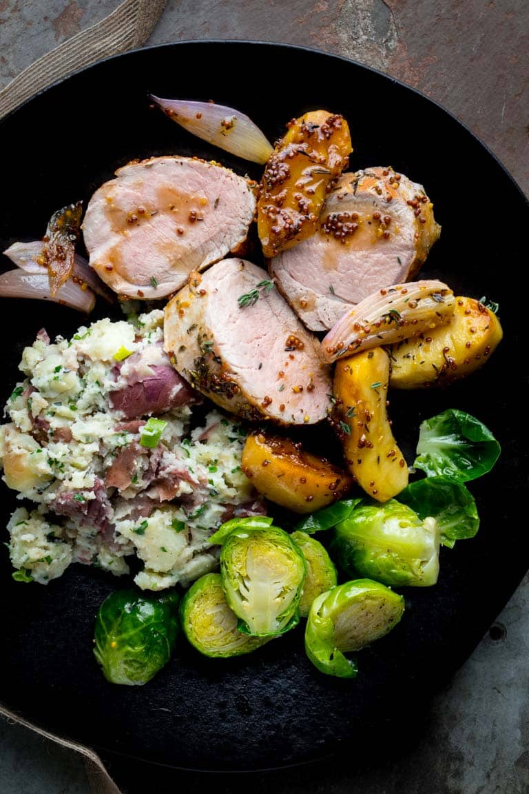 A black plate with pork tenderloin, herb smashed potatoes and brussels sprouts on a slate surface