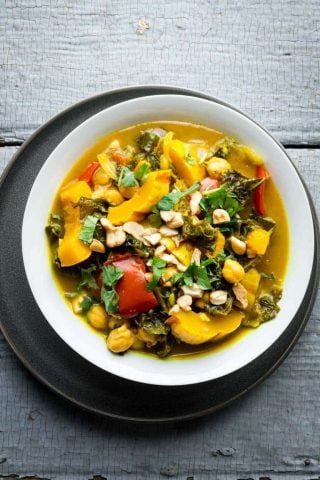 An overhead view of a white bowl filled with vegetable curry topped with cilantro and cashews on a gray painted surface