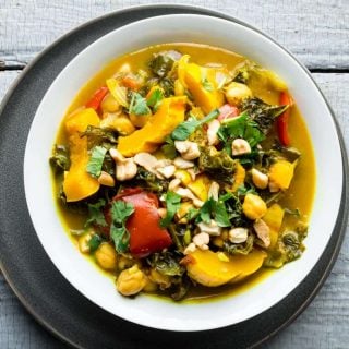 An overhead view of a white bowl filled with vegetable curry topped with cilantro and cashews on a gray painted surface