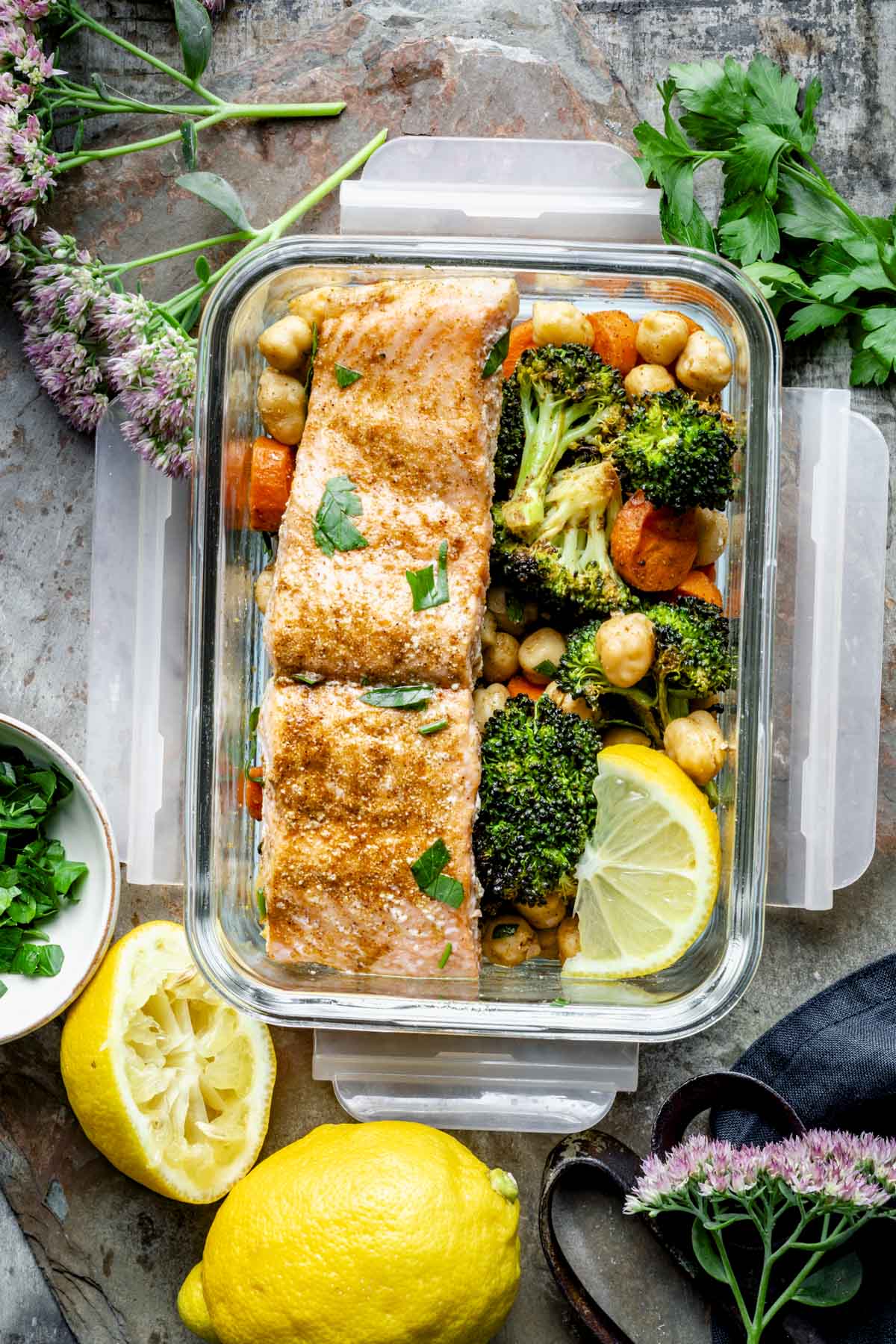 a meal prep container filled with the moroccan spiced salmon filet, some of the veggies and chickpeas and lemons on the side