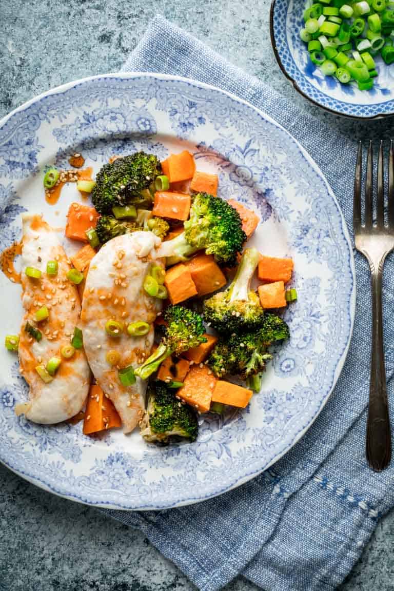 A blue and white plate with Chicken tenders cooked with broccoli and sweet potatoes with honey sriracha sauce on top.
