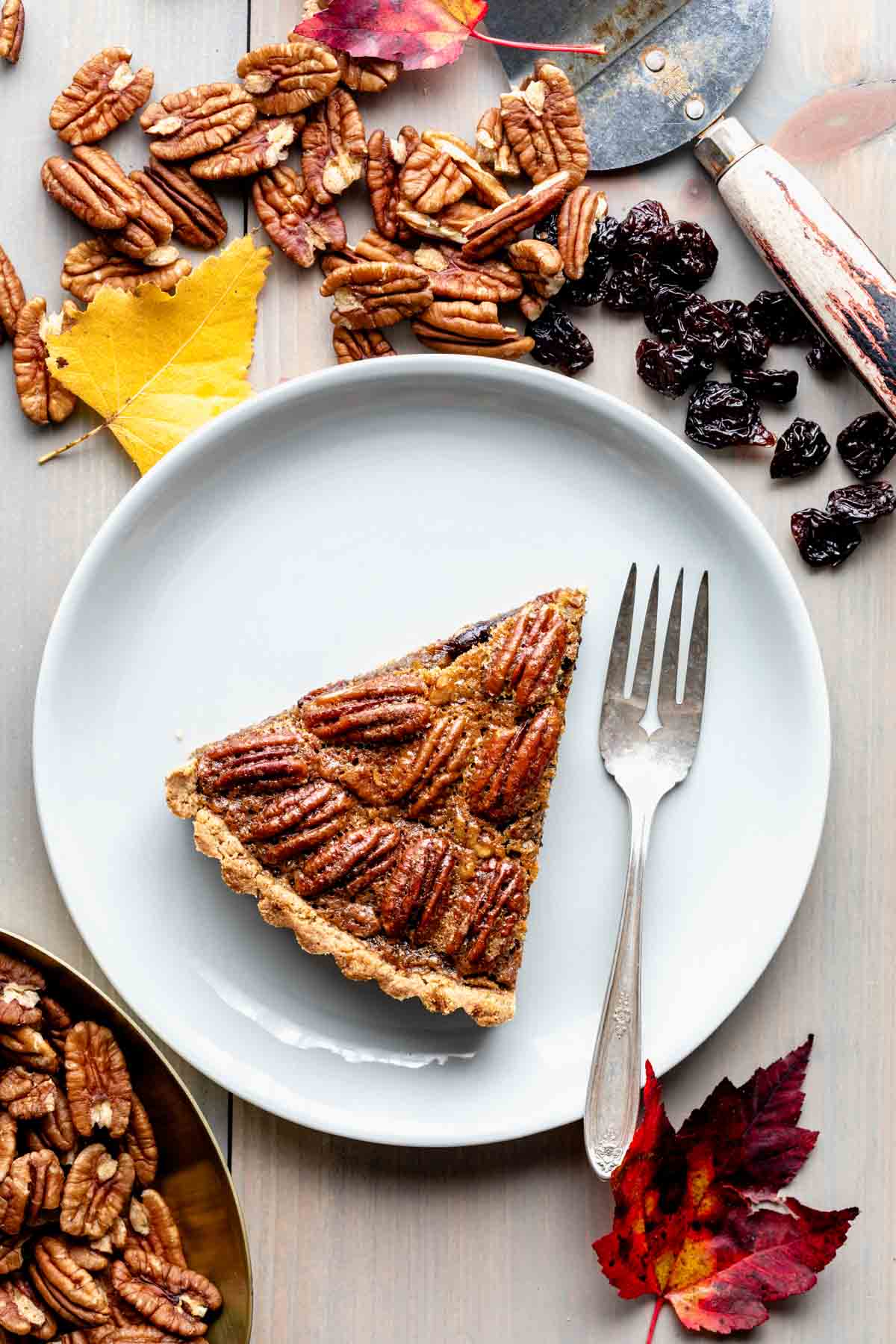 a slice of pecan tart on a table with foliage, nuts and dried cherries