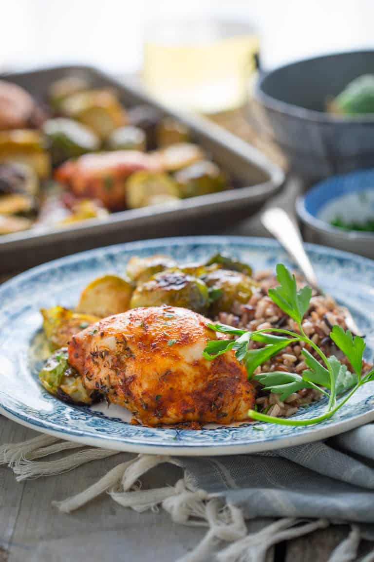 Side view of blue plate with chicken and brussels sprouts, a sheet pan and glass of wine in the background