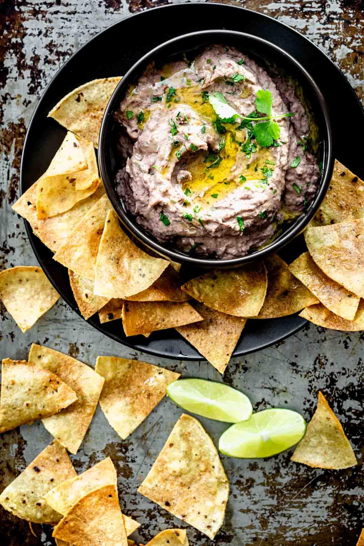 black bowl of black bean hummus with tortilla chips on a black platter with lime wedges on a metal sheet pan