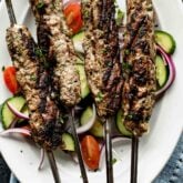 four kofta kebabs on a platter with cucumbers and tomatoes