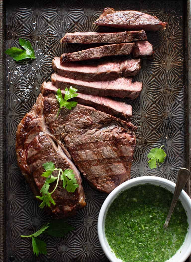 a sliced steak on a black baking sheet with a bowl of chimichurri sauce