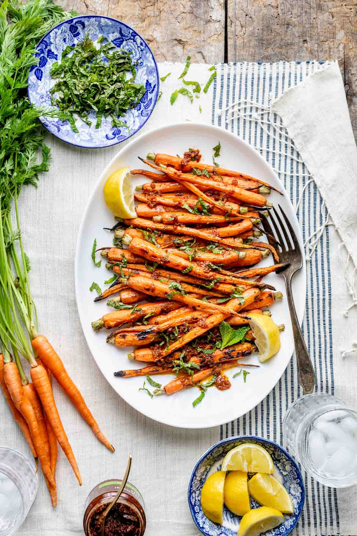 An overhead photo of a farm table with carrots with tops, a platter of grilled carrots and a dish of chopped mint.