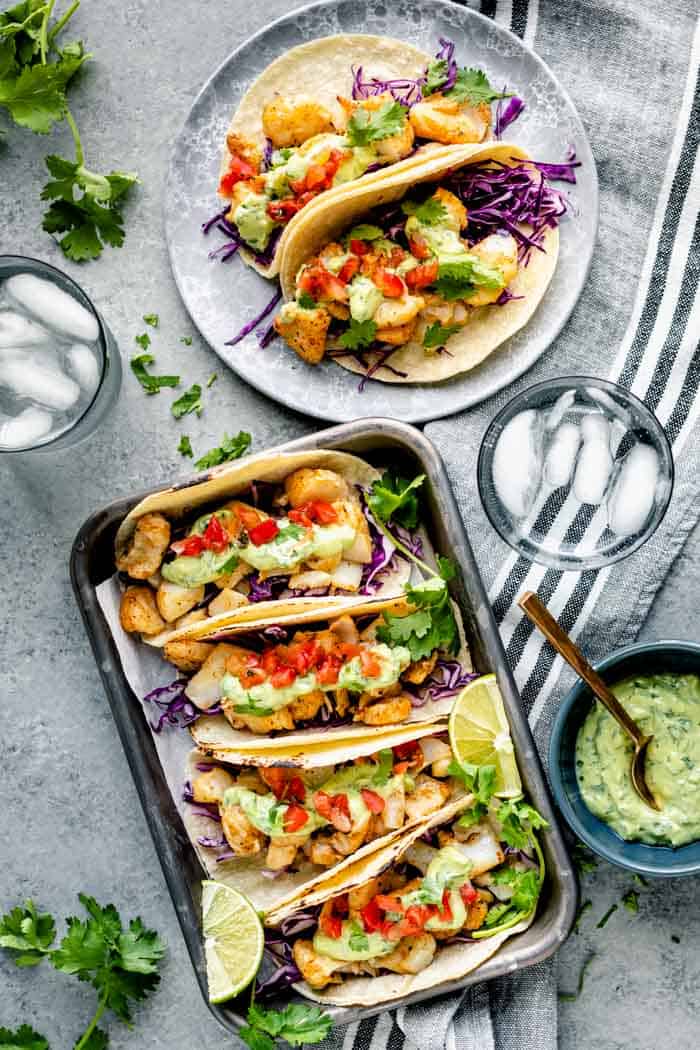 An overhead view of a table with a tray of cod fish tacos and a plate with two tacos on it.