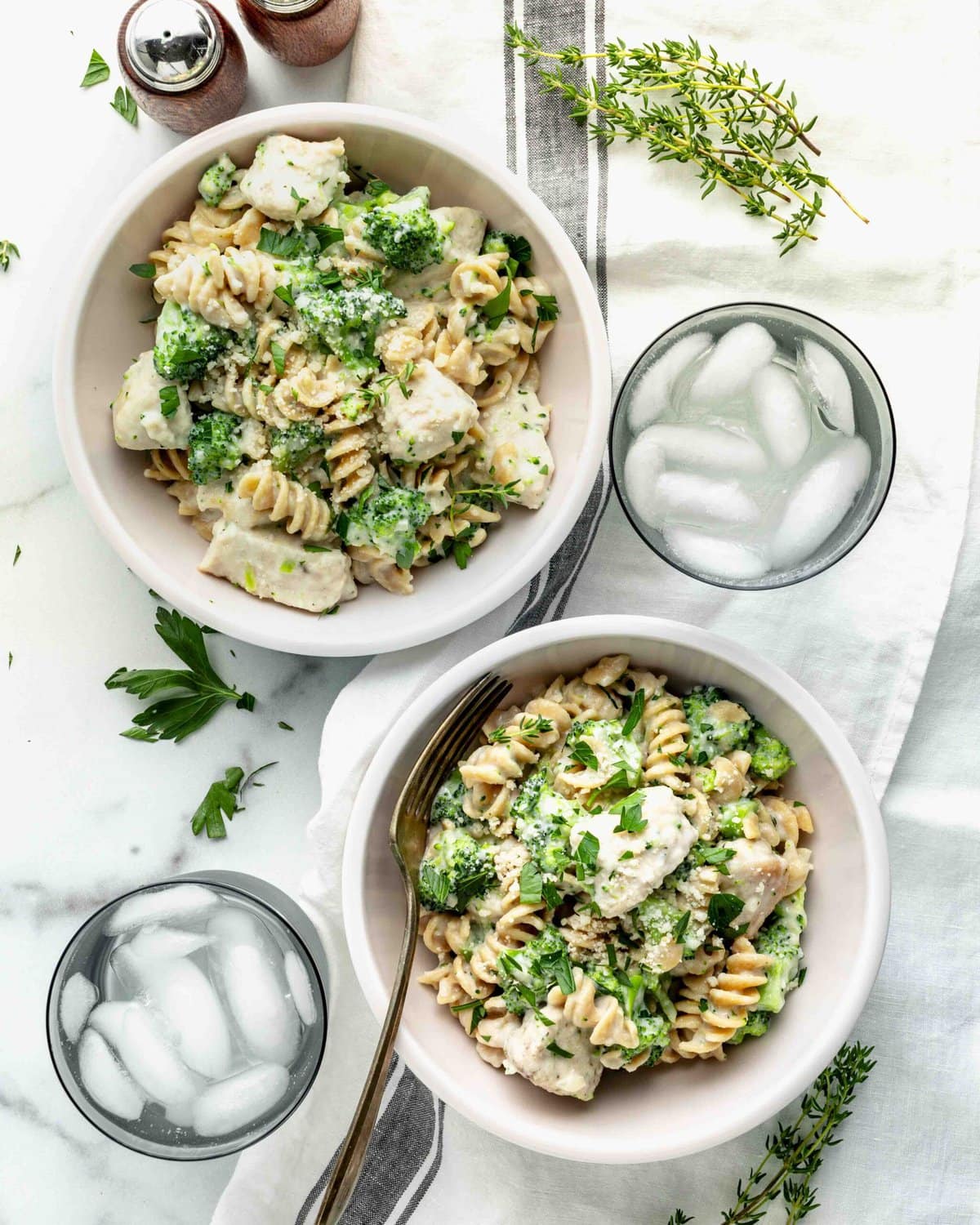 A marble counter with two bowls of chicken alfredo with broccoli plus two glasses of ice water