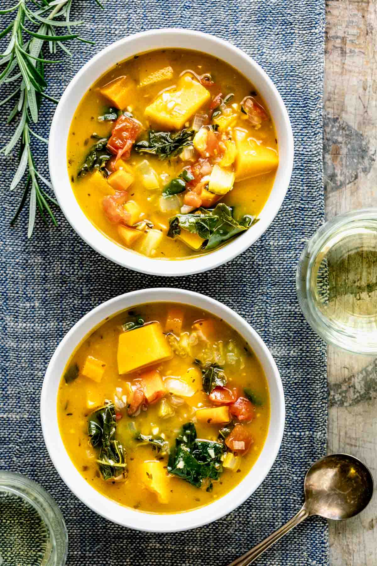 two white bowls of veggie soup with rosemary sprigs on a blue placemat