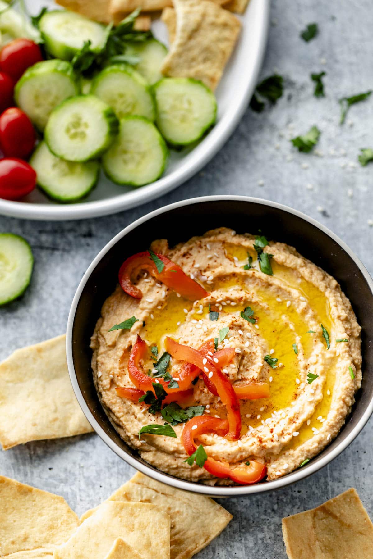 the hummus in a bowl on a bluish gray concrete surface with fresh veggies and pita chips