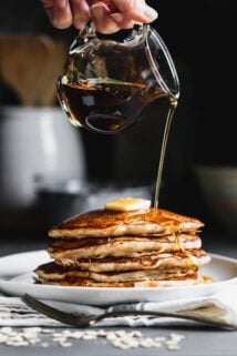 Oatmeal Pancakes with a pitcher or syrup pouring over a stack of pancakes