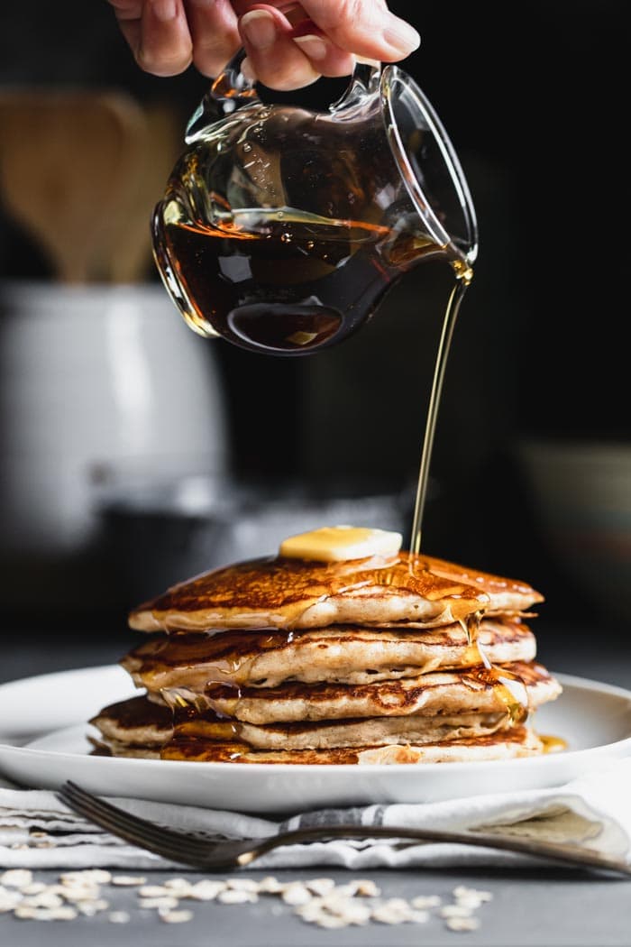 Oatmeal Pancakes with a pitcher or syrup pouring over a stack of pancakes