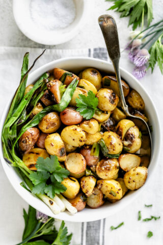 a white bowl with grilled potatoes in it with a couple of grilled scallions on the side and chive blossoms on the white table