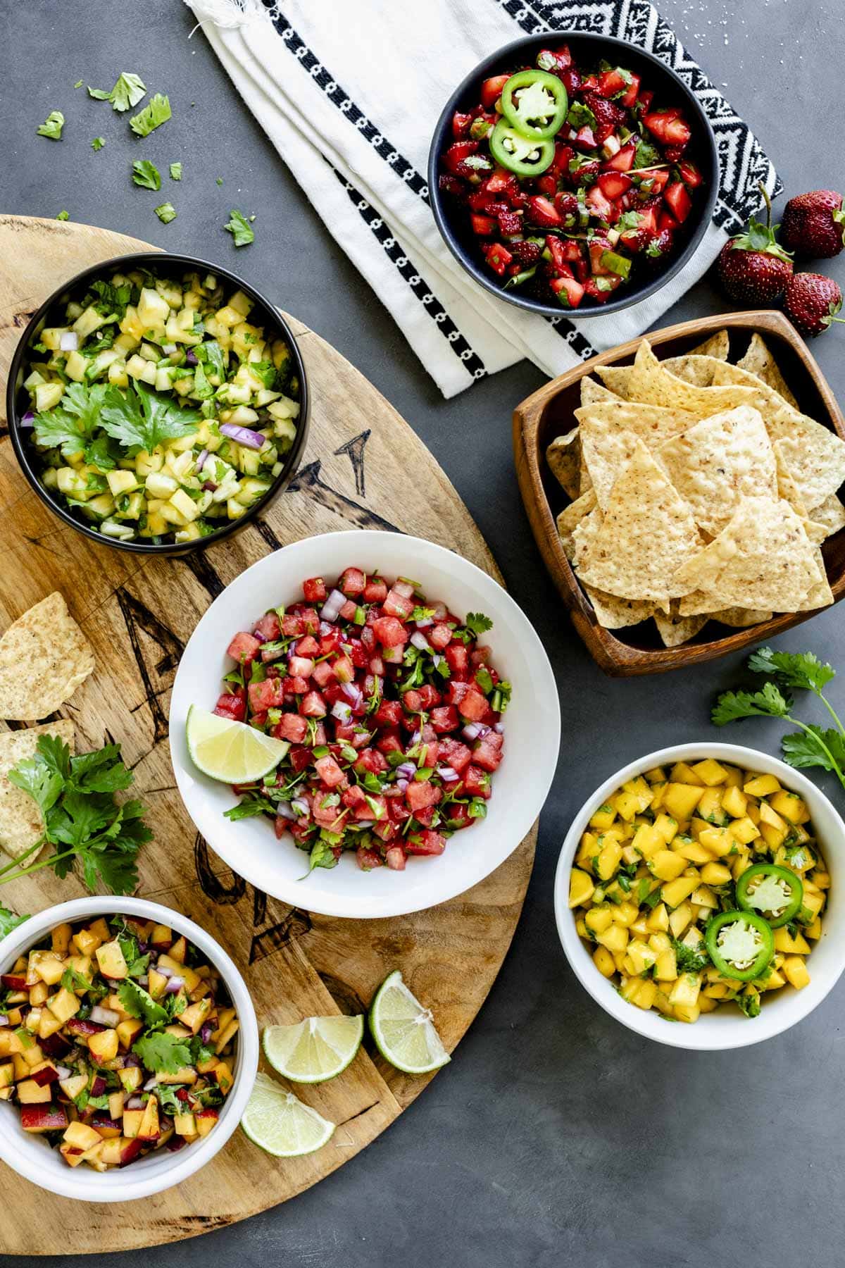 several bowls of the fruit salsa on a table top with a wooden board, chips and a white and black napkin