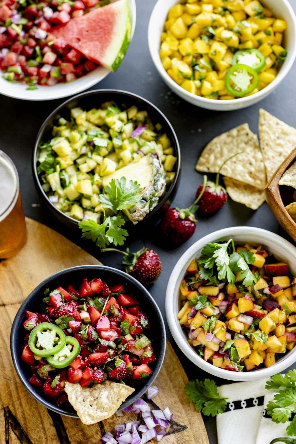 several bowls of the fruit salsa on a table top with a wooden board, chips and a beer