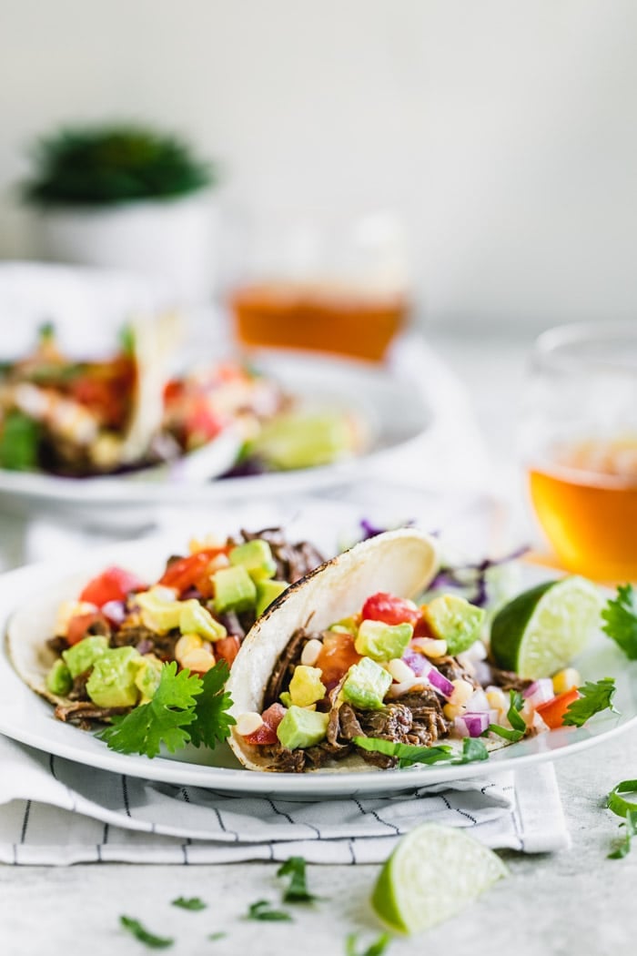 Two beef barbacoa tacos on a white plate in the foreground with another plate of tacos and glasses of beer in the background.