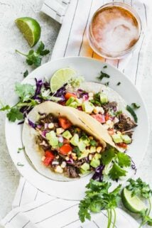 Two beef barbacoa tacos on a white plate on top of a linen with a beer, limes and cilantro sprigs.