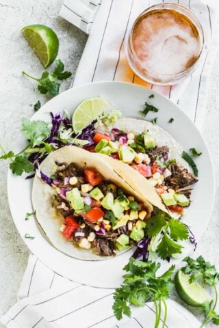 Two beef barbacoa tacos on a white plate on top of a linen with a beer, limes and cilantro sprigs.