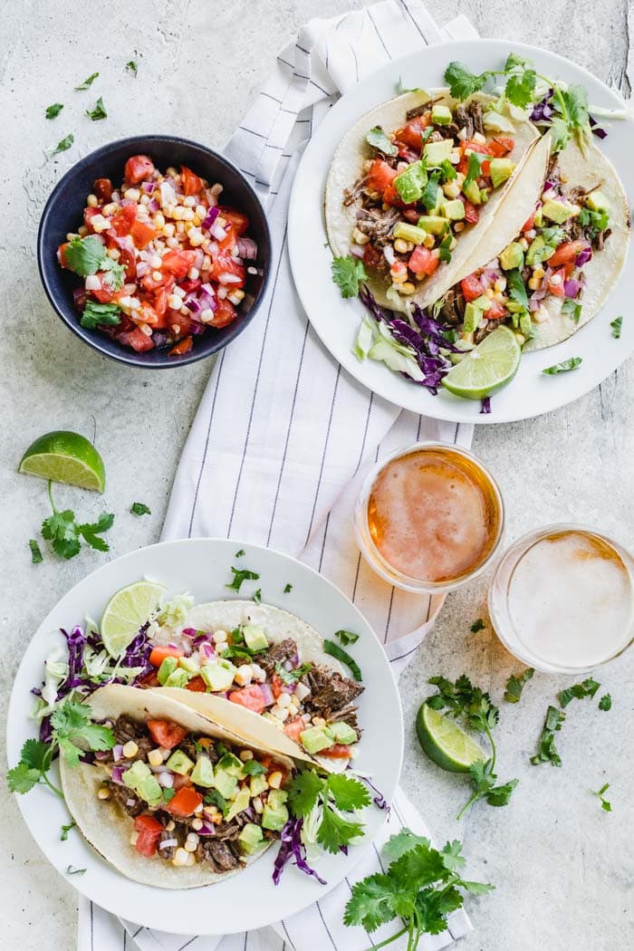 Overhead shot of two white plates on top of a linen with two beef barbacoa tacos on each plate and 2 glasses of beer and a bowl of corn salsa.