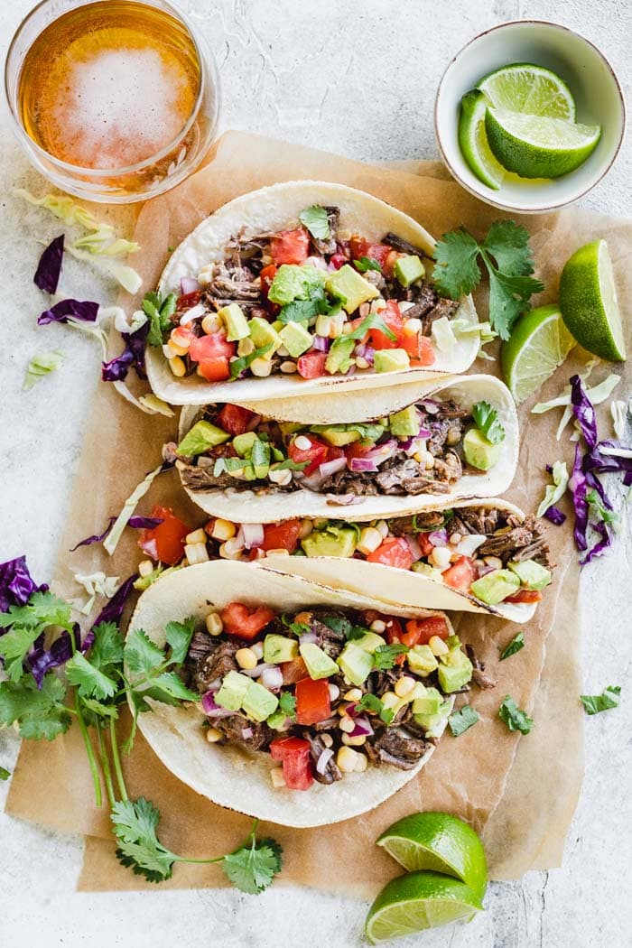Four beef barbacoa tacos topped with a corn salsa on to of brown parchment and surrounded by lime slices, cilantro sprigs and a glass of beef.