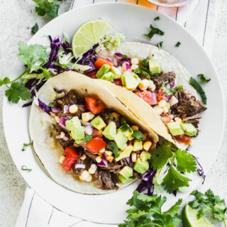 Overhead shot of two beef barbacoa tacos topped with corn salsa on a white plate on top of a linen.