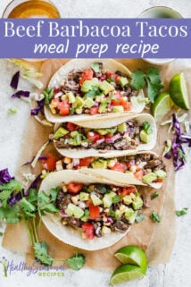 Four beef barbacoa tacos topped with a corn salsa on to of brown parchment and surrounded by lime slices, cilantro sprigs and a glass of beef.