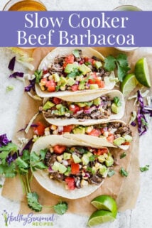 Four beef barbacoa tacos topped with a corn salsa on to of brown parchment and surrounded by lime slices, cilantro sprigs and a glass of beef.