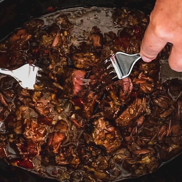 Hands using two forks to shred the beef into the sauce in the slow cooker.
