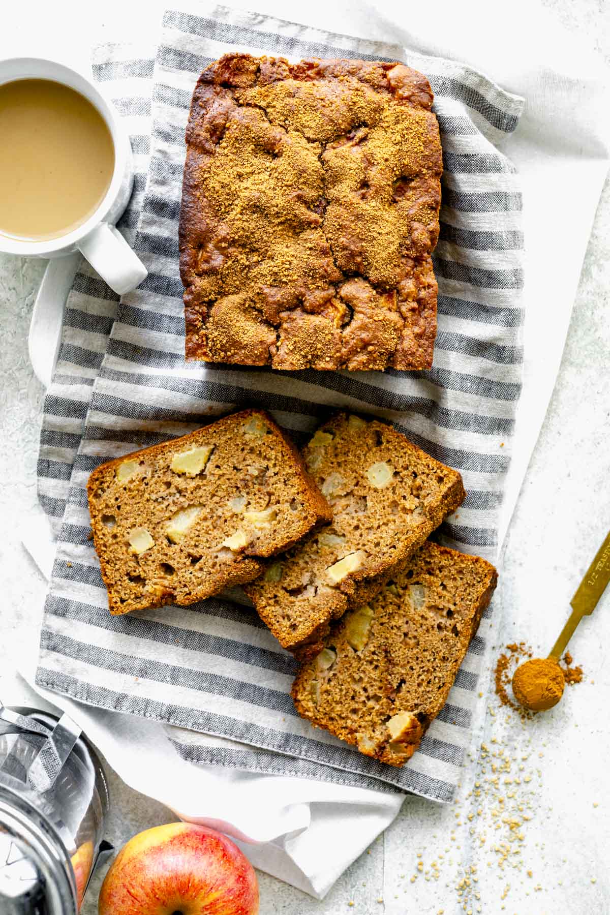 the loaf of apple bread on a white and gray striped towel with a foamy coffee and apple with a little spoonful of cinnamon
