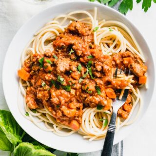 Overhead shot of white bowl filled with slow cooker bolognese and pasta and fresh herbs on a linen