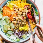overhead of a turquoise painted salad bowl with garden salad in it, two salad tongs and hands on the tongs