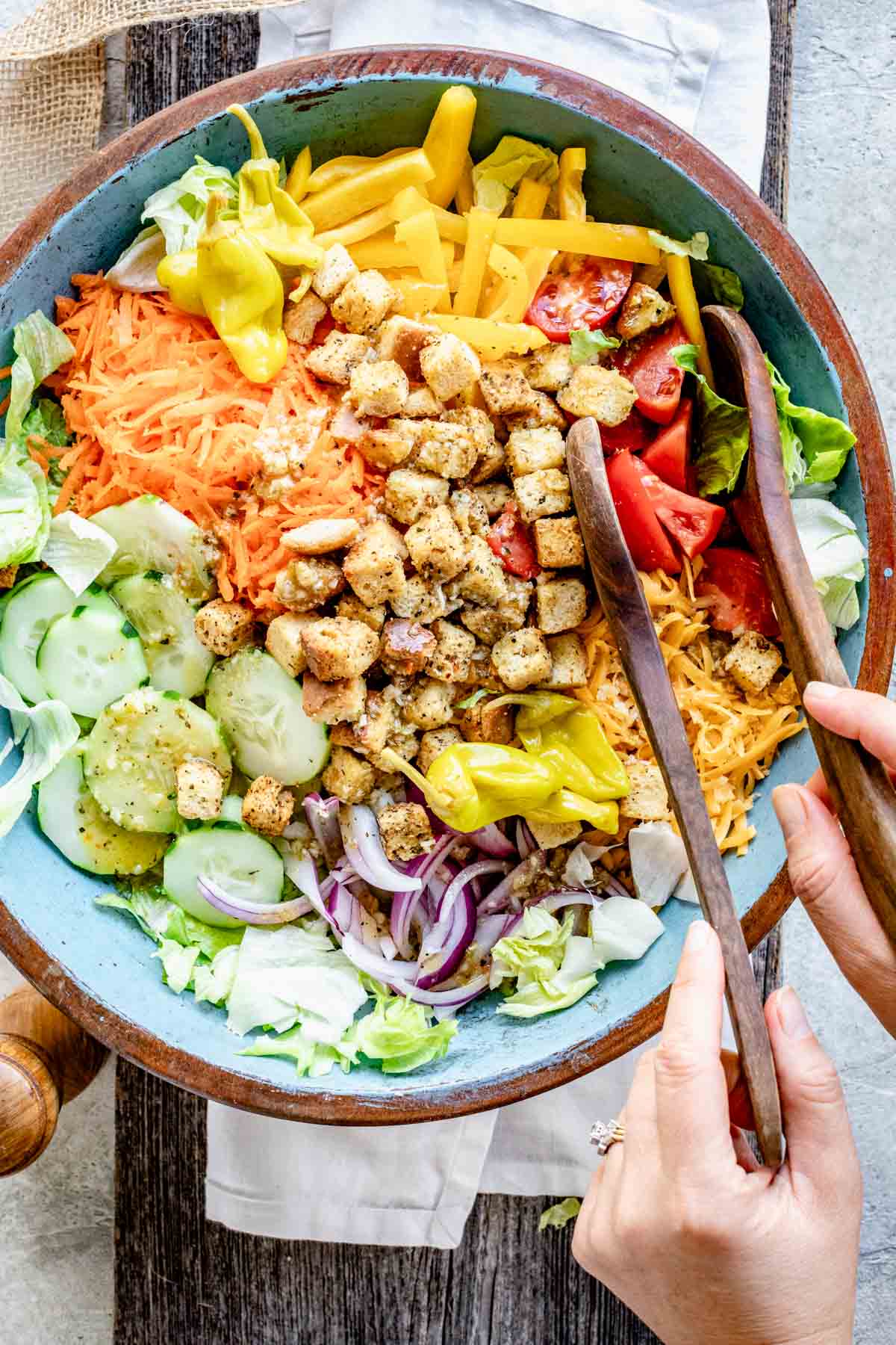 overhead of a turquoise painted salad bowl with garden salad in it, two salad tongs and hands on the tongs