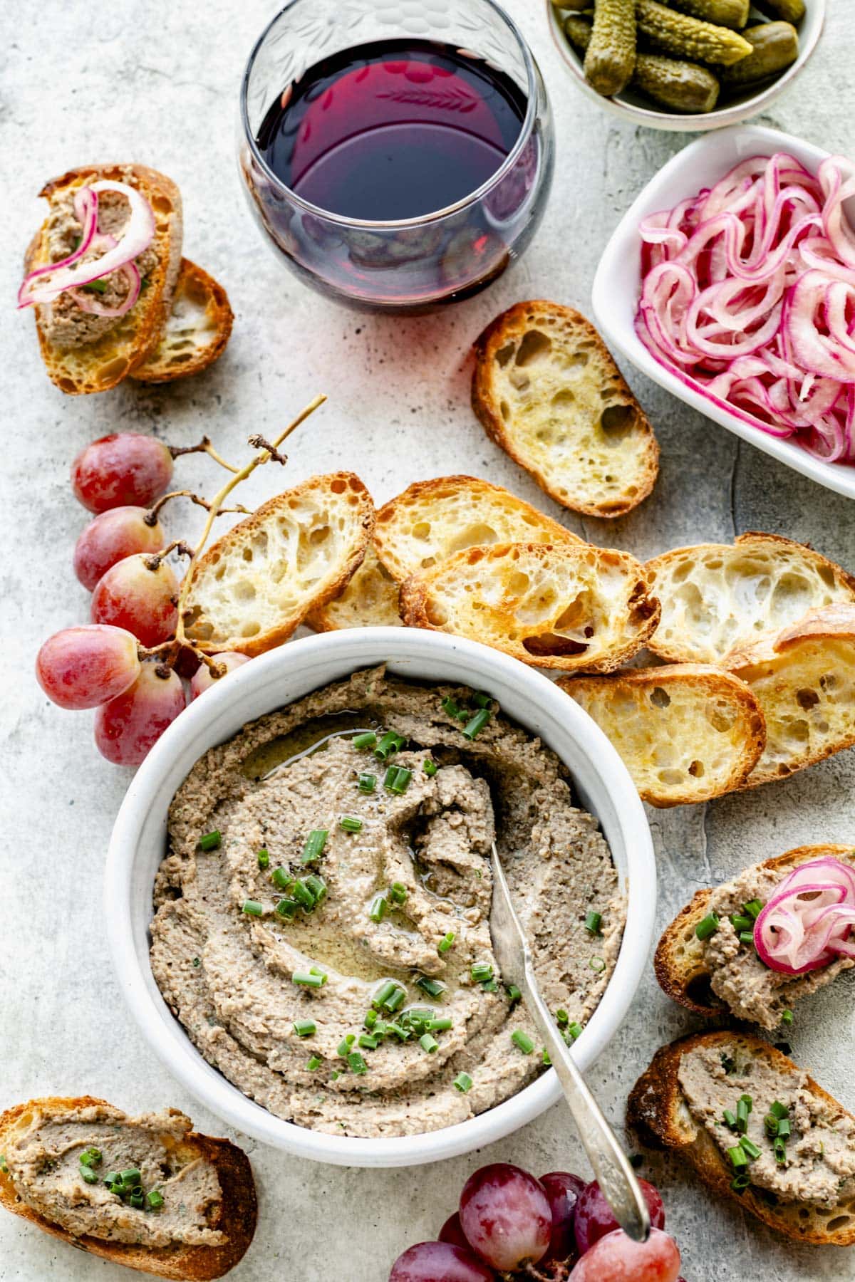 the pate in a ceramic crock set on a white table with grapes, crostini, pickled onions and a glass of red wine