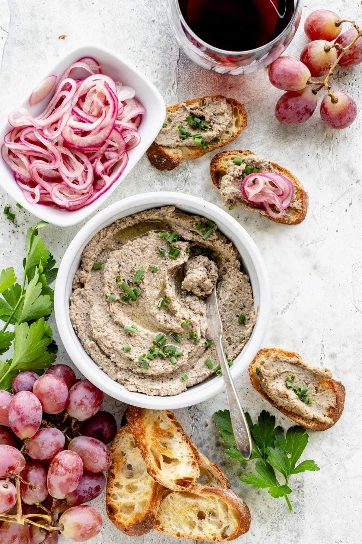 the pate in a ceramic crock set on a white table with grapes, crostini, pickled onions and a glass of red wine