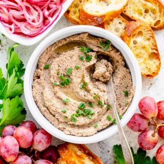 the pate in a ceramic crock set on a white table with grapes, crostini, pickled onions and a glass of red wine