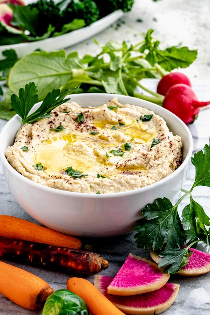Cauliflower Hummus in a bowl from the side with some veggies on a marble surface