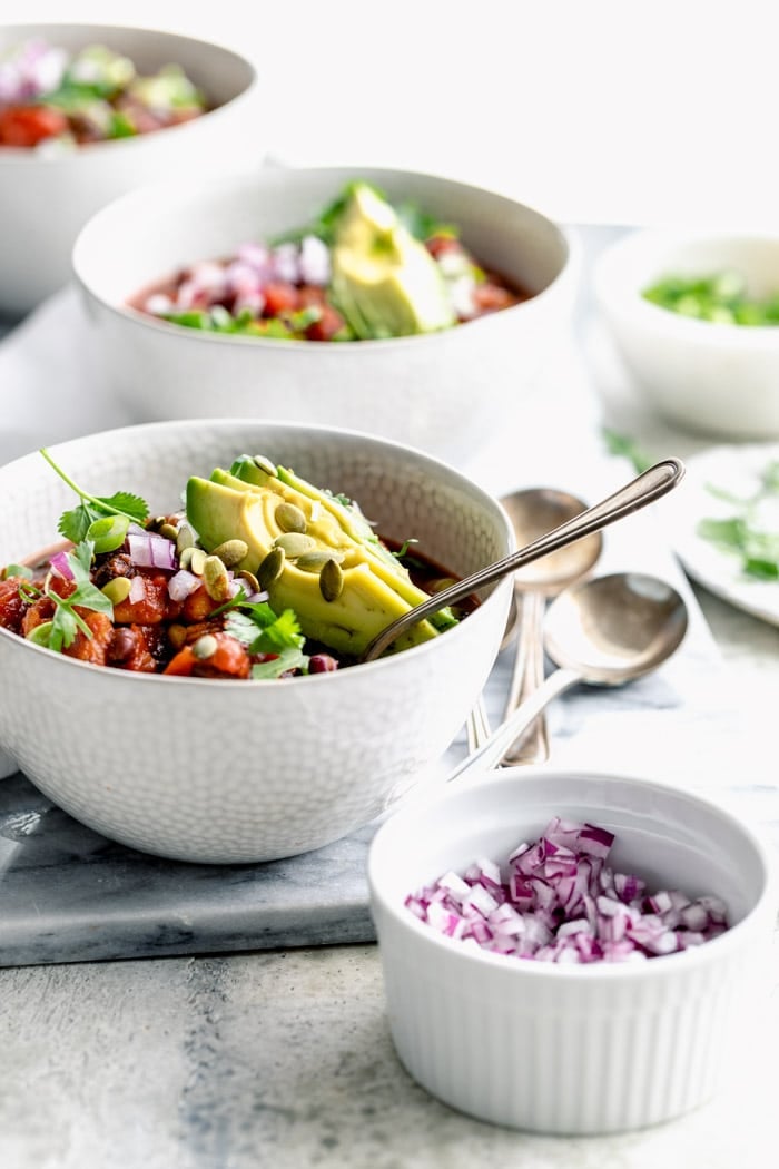 bowls of veggie chili on a white table with white light from behind. Dishes of toppings in the foreground