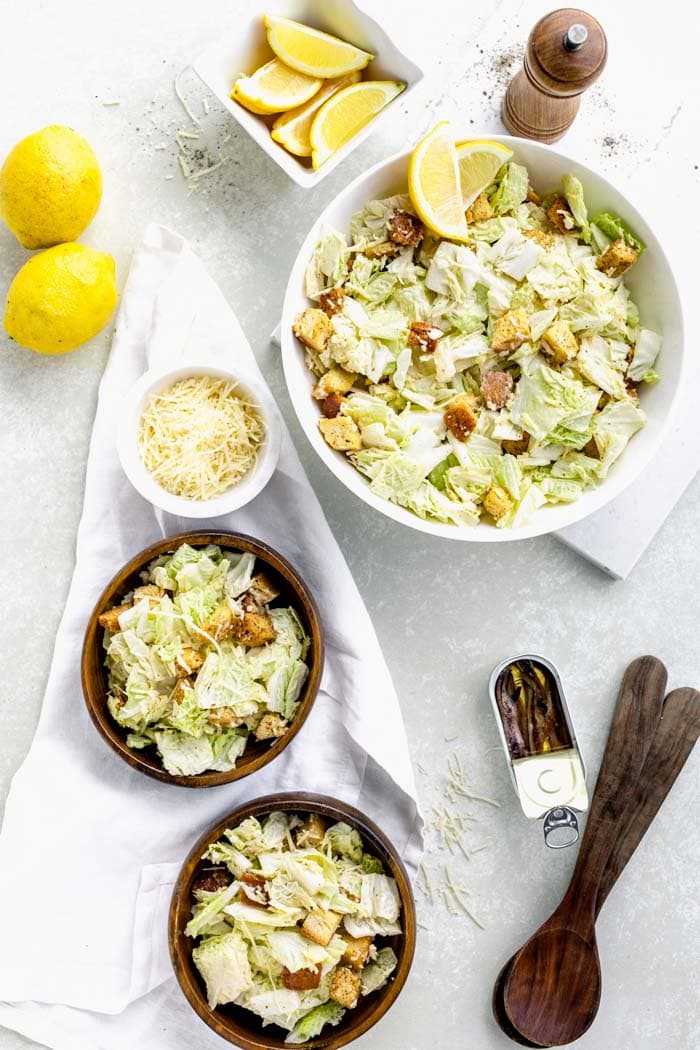 overhead tablescape with bowls of Caesar, anchovies in a tin, lemons and tongs
