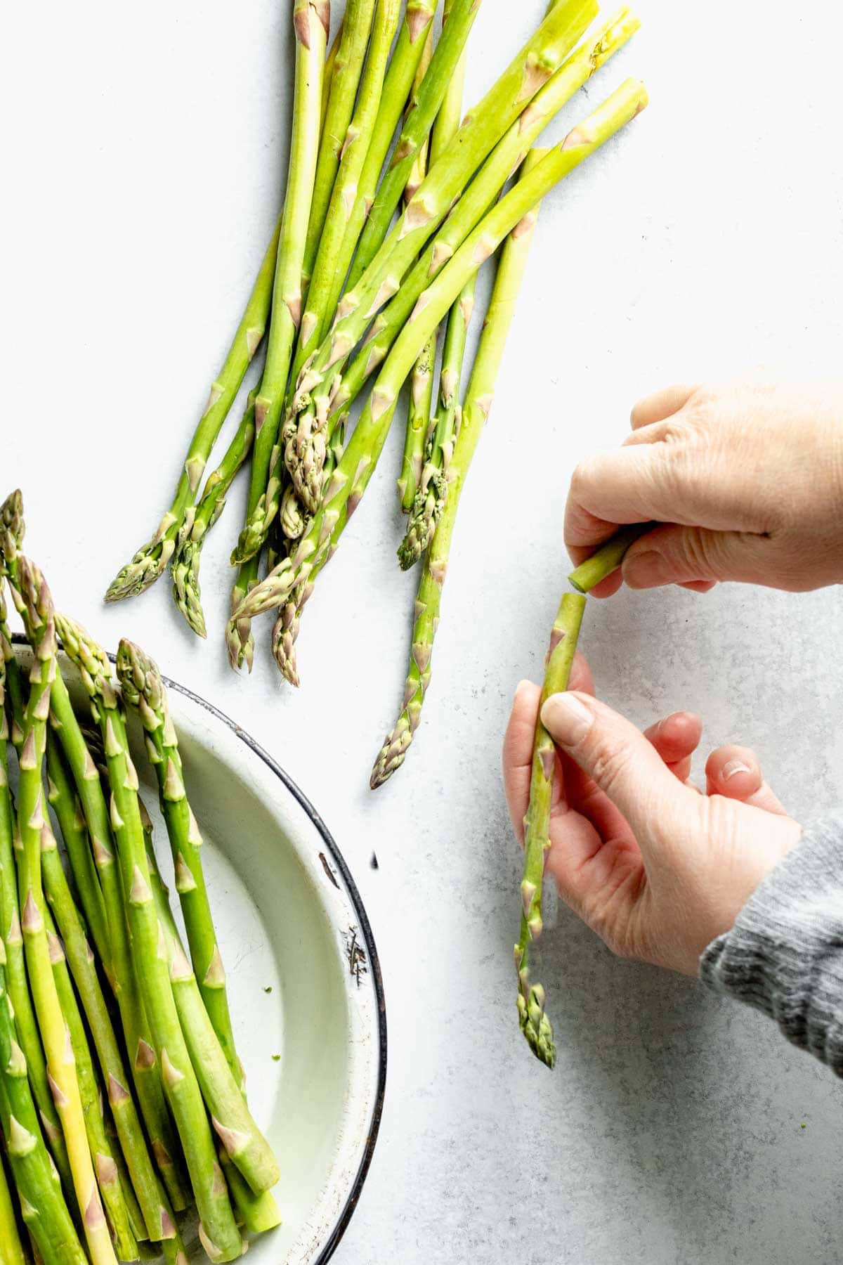 Hands holding a piece of asparagus, a bowl of asparagus sits nearby.