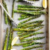 roasted asparagus on a sheet pan with tongs, herbs and lemon wedges