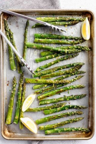 roasted asparagus on a sheet pan with tongs, herbs and lemon wedges