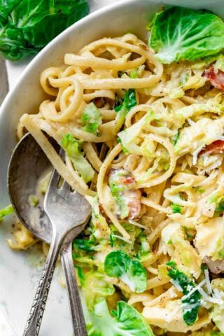 the pasta in a white bowl, close up with creamy sauce, bits of bacon and brussels sprouts leaves sprinkled over the serving for garnish