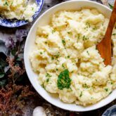 Close up overhead of the bowl of mashed potatoes, a serving in a blue bowl and foliage on the table.
