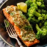 Salmon filet topped with creamy herb sauce set on a plate with broccoli, lemon wedge and a fork.