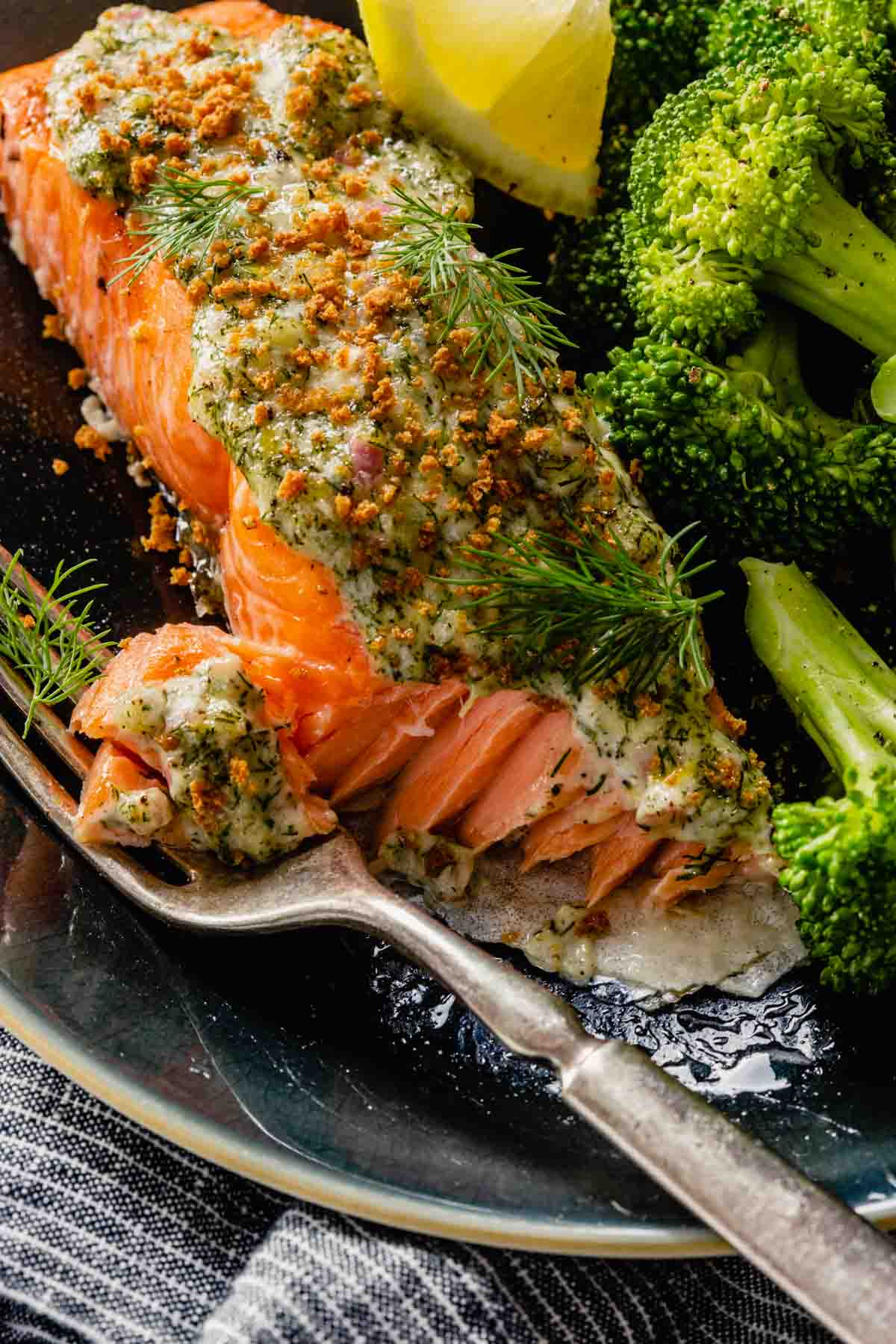 Salmon filet topped with creamy herb sauce set on a plate with broccoli, lemon wedge and a fork.