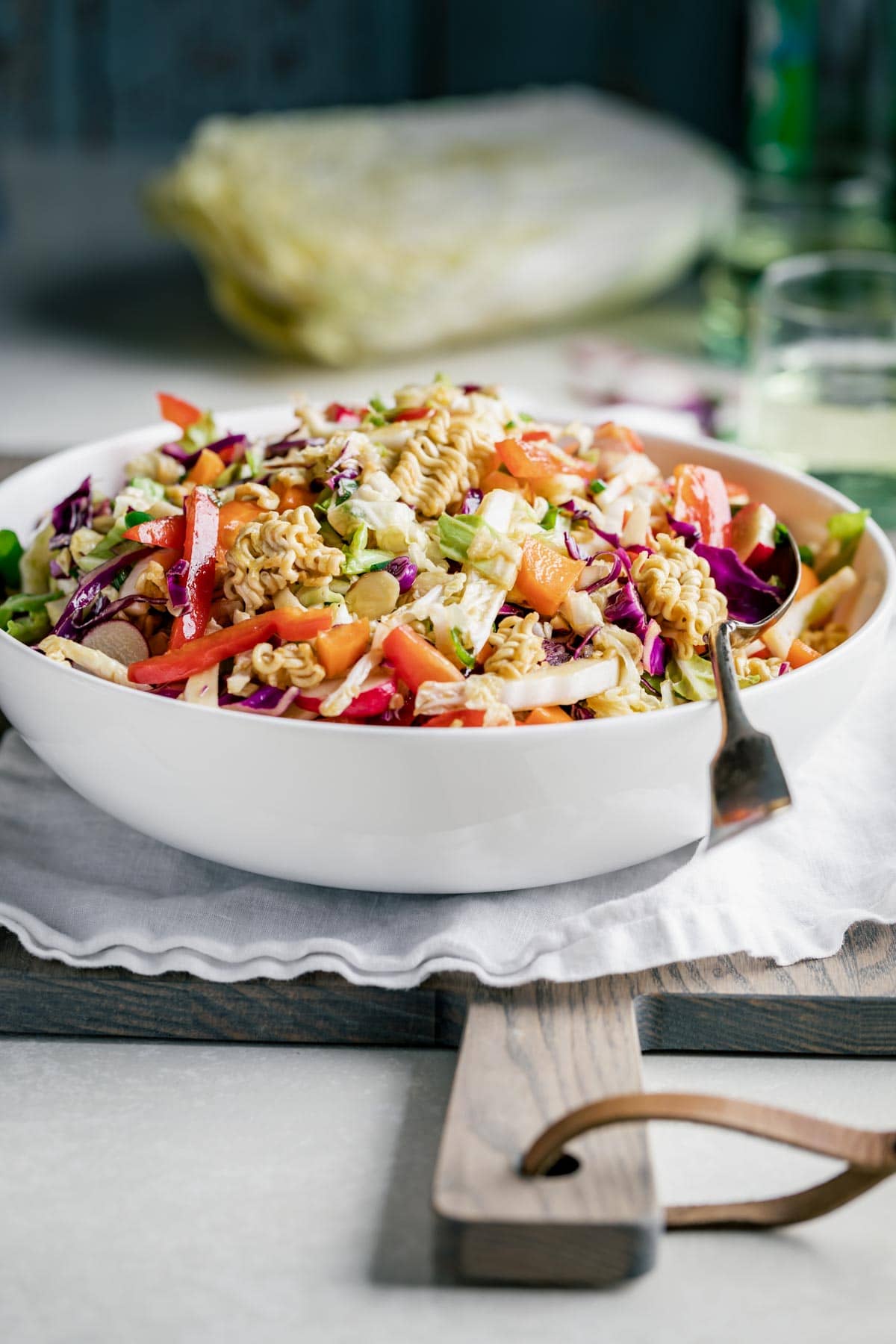 Napa Cabbage Ramen Noodle Salad in a white bowl with a silver spoon in it, on a chopping board on a table.