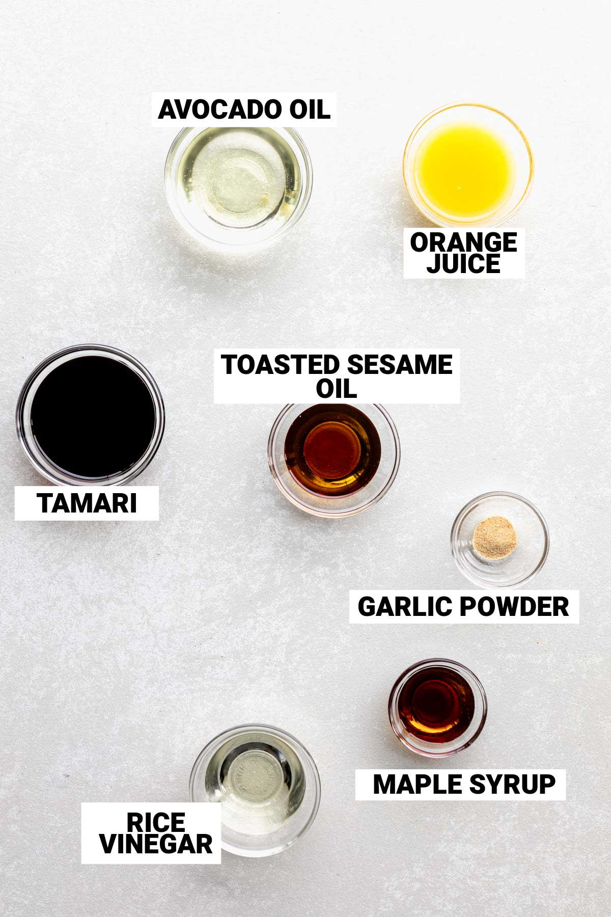 Top view of avocado oil, soy sauce, rice vinegar, orange juice, sesame oil, maple syrup, and garlic powder each in small bowls on a white table with text labels on them.