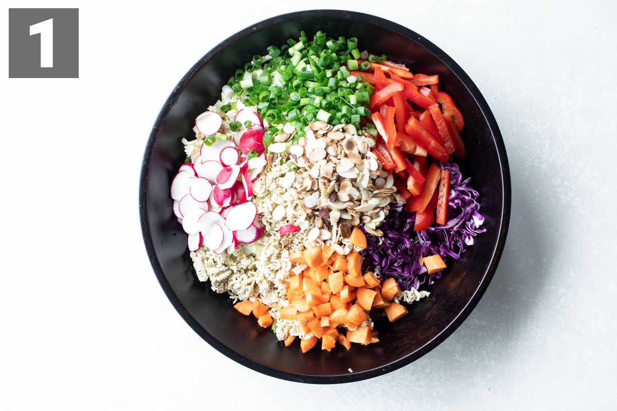 Top view of napa Cabbahe Ramen Noodle Salad ingredients separated into kinds in a black bowl.