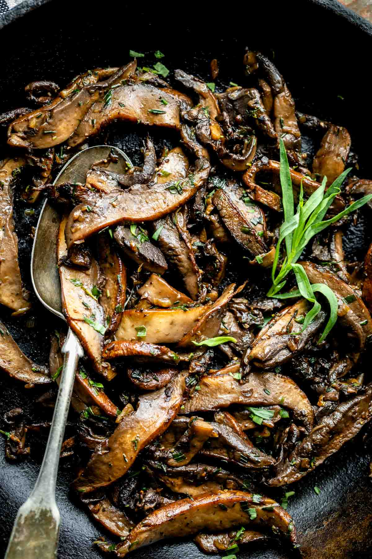 Top view of a close up of sautéed portobello mshrooms in a skillet with a spoon in it and fresh herb leaves on top.